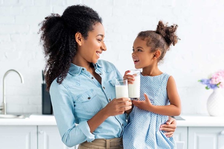smiling african american mother and daughter holding glasses of milk in kitchen and looking at each other