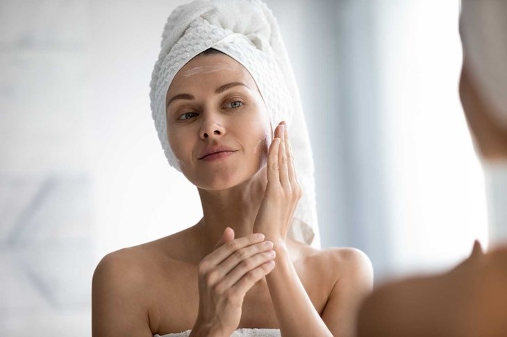 Head shot close up beautiful peaceful woman applying moisturizer on skin, looking in mirror, standing in bathroom, pretty young female with white bath towel on head using cream, touching face